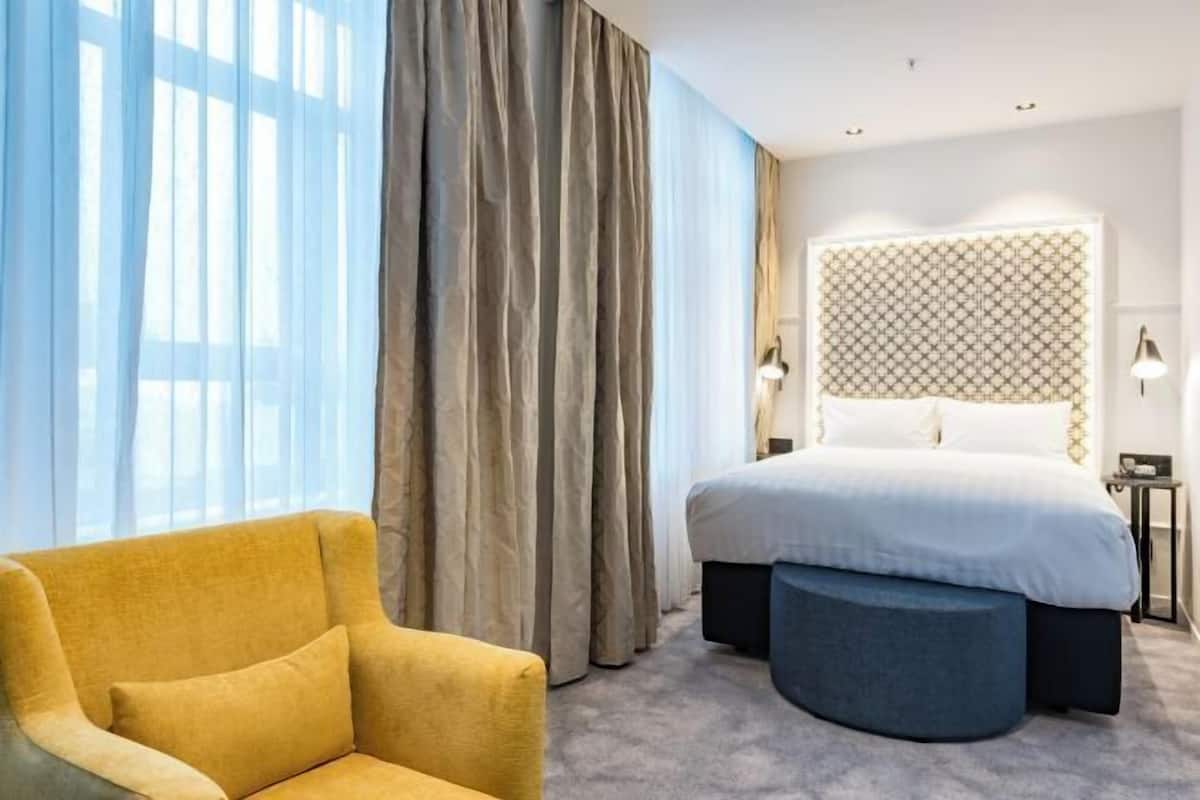 A hotel room with a bed, a chair, a round ottoman, curtains, and a patterned headboard.