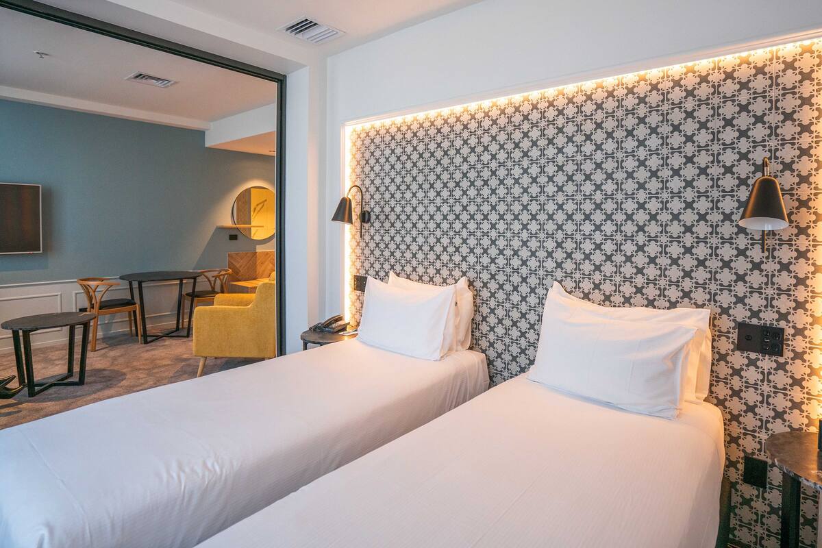 A hotel room with two beds, a desk, a chair, a lamp, and a clock on the nightstand.