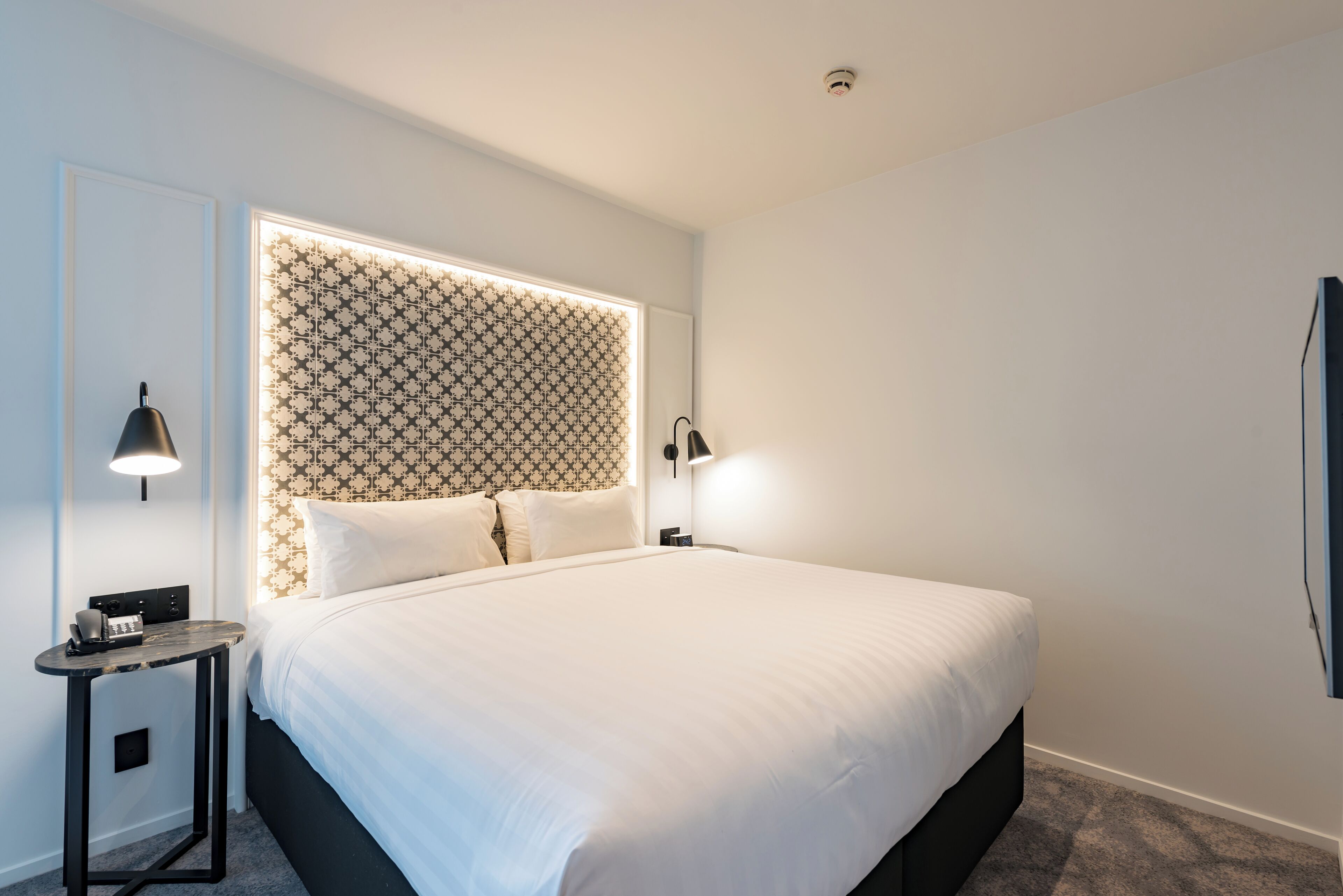 A neatly made bed with a patterned headboard, a bedside table with a lamp, and a wall-mounted TV.