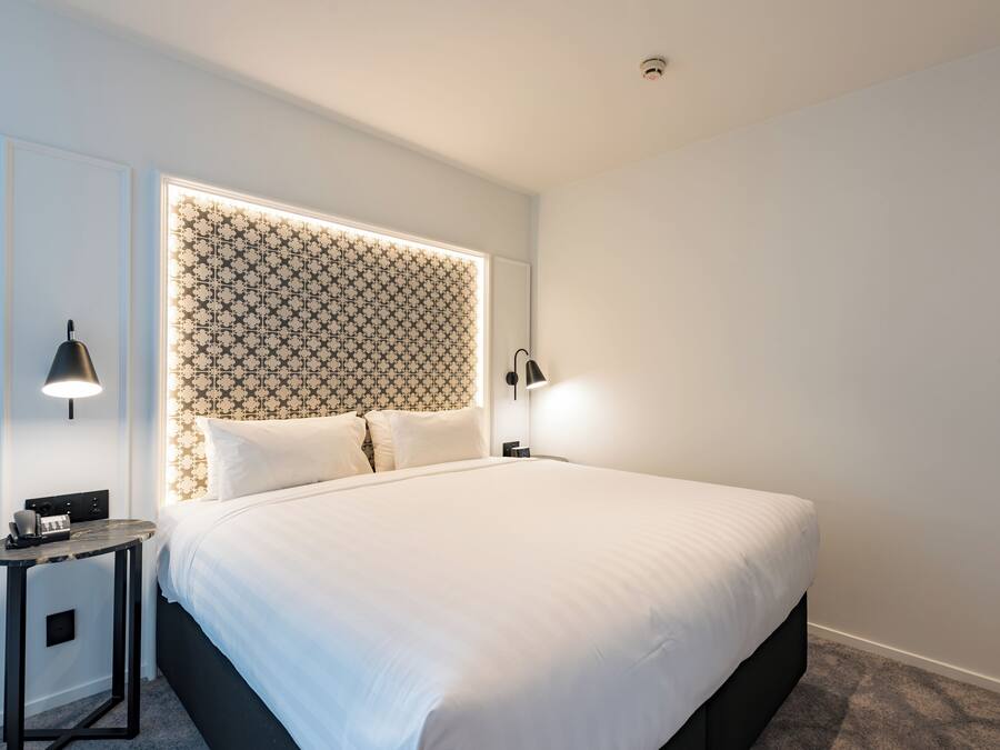 A neatly made bed with a patterned headboard, a bedside table with a lamp, and a wall-mounted TV.