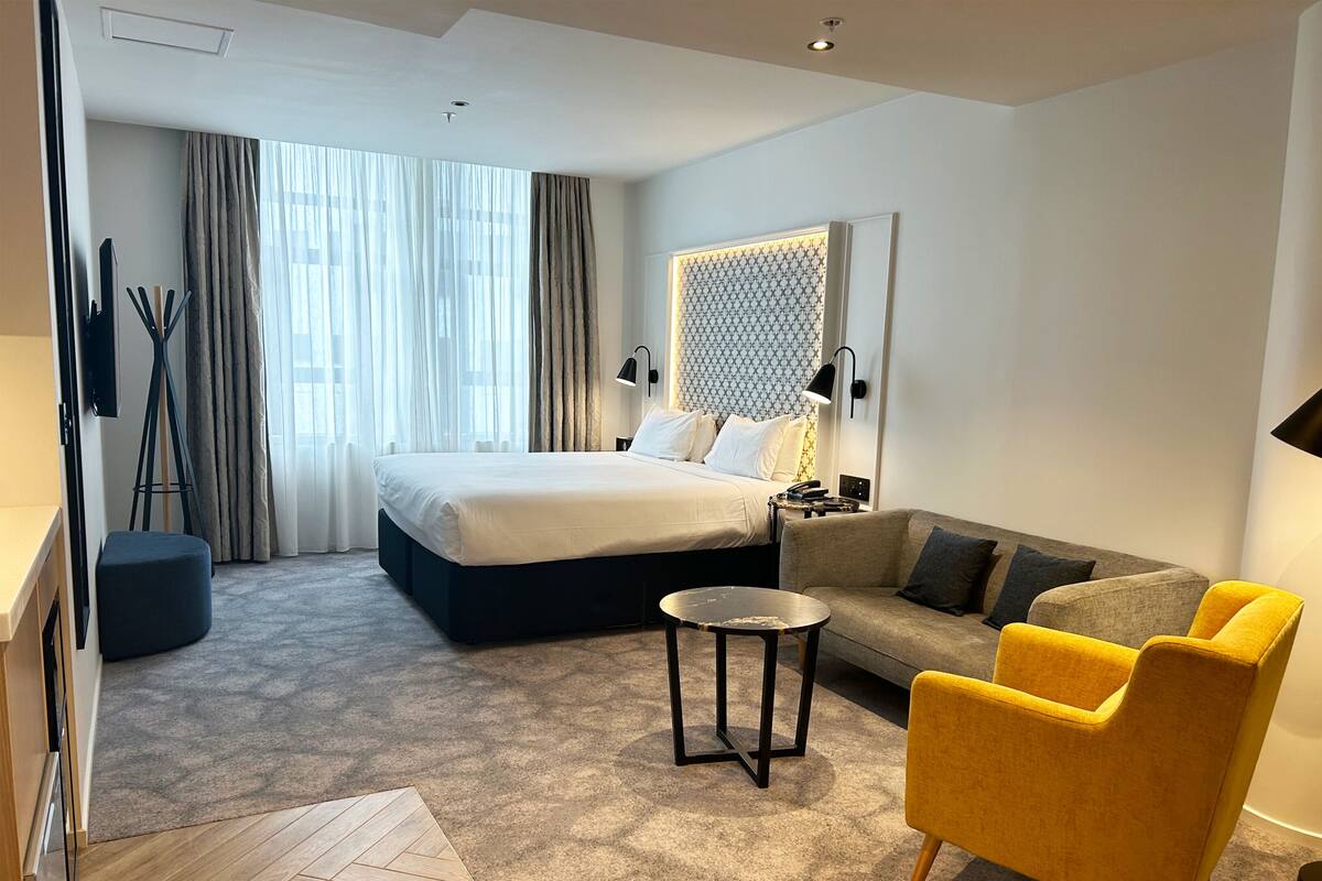 A hotel room with a bed, a sofa, a yellow armchair, a small table, and a patterned wall.