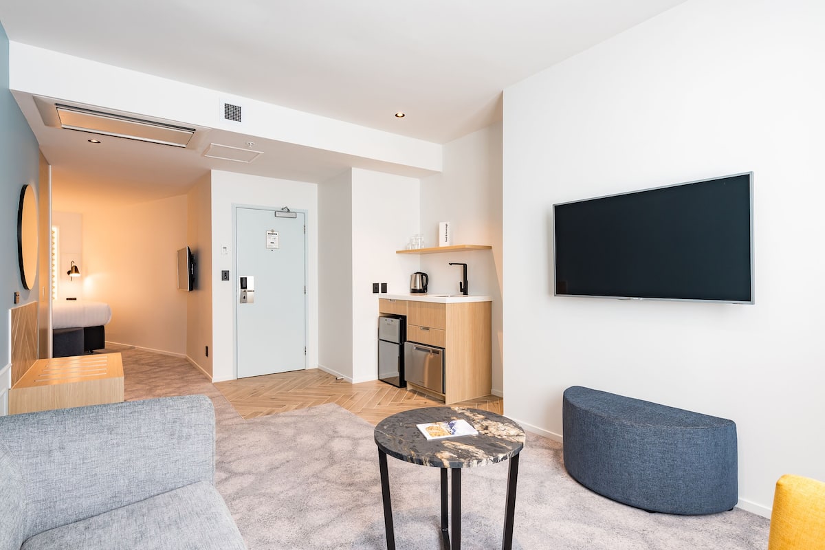 A modern hotel room with a grey sofa, a round coffee table, a blue ottoman, a flat-screen TV mounted on the wall, a kitchenette with a fridge and microwave, and a wooden door leading to a bathroom.