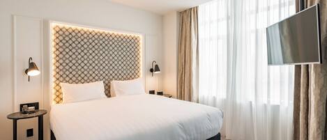 A modern hotel room with a large bed, a headboard with a patterned design, two bedside tables with lamps, a flat-screen TV, and sheer curtains.