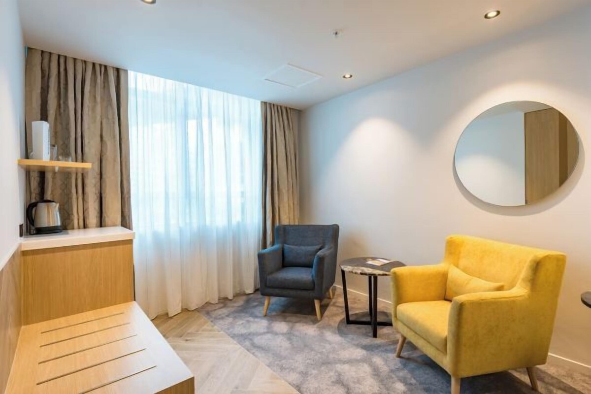 Habitación de hotel con cama, una mesita, dos sillas, un espejo redondo y una ventana con cortinas.
