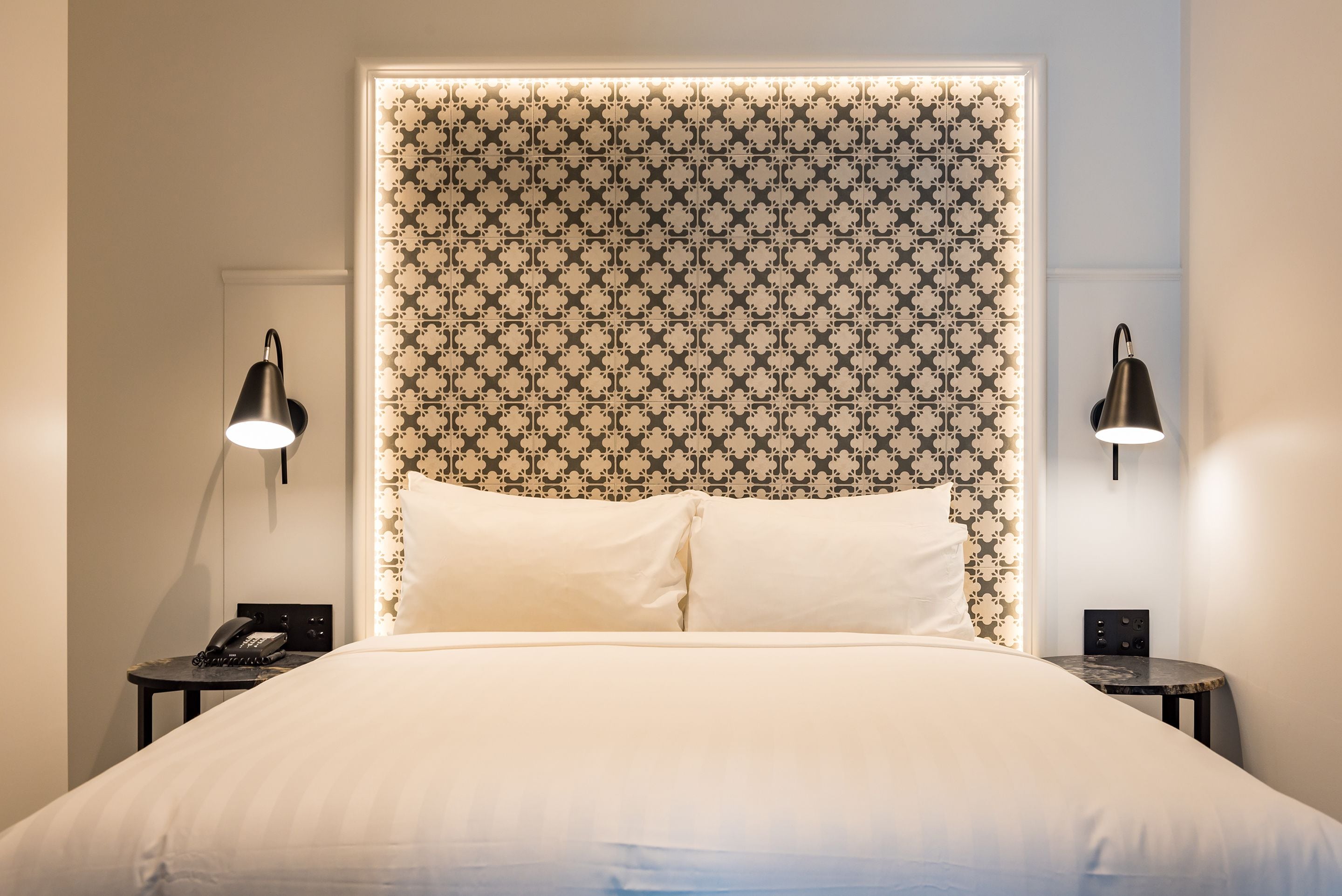 A neatly made bed with a patterned headboard, two bedside lamps, and a small table with a phone on each side.