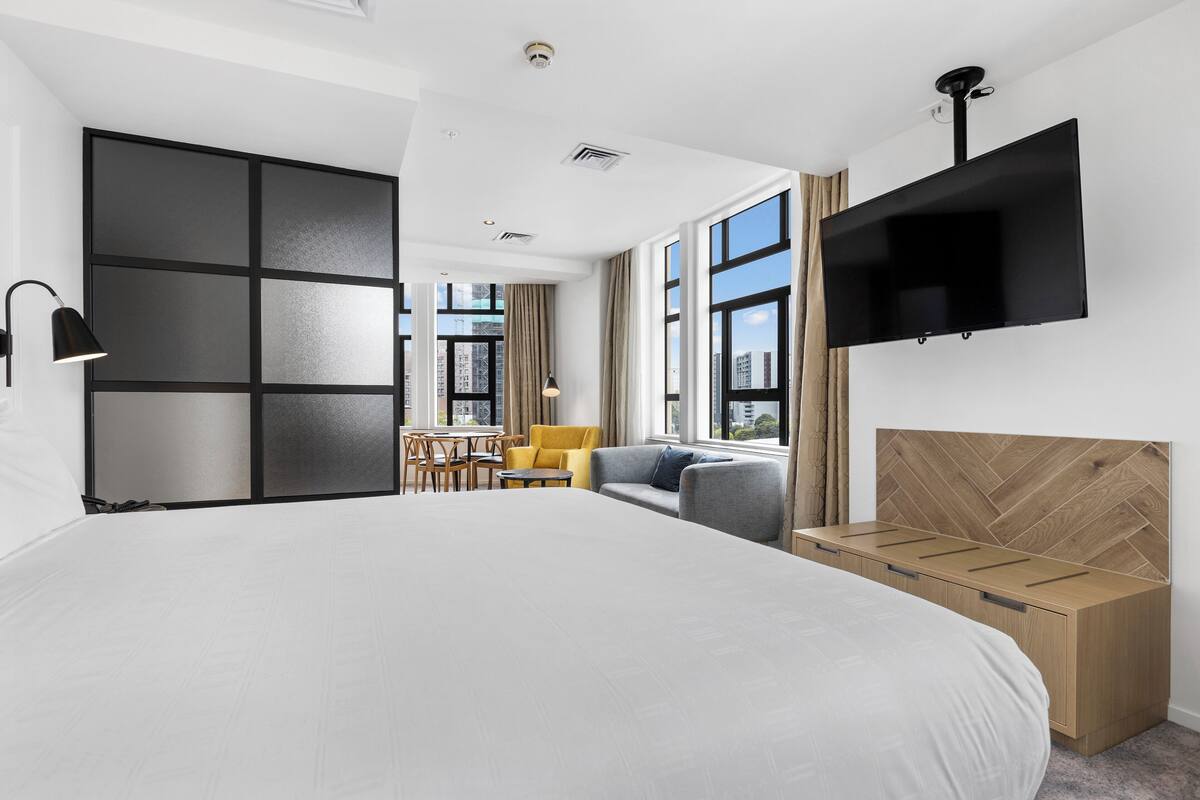 A modern hotel room with a large bed, a flat-screen TV, a wooden headboard, a desk, and a sofa.