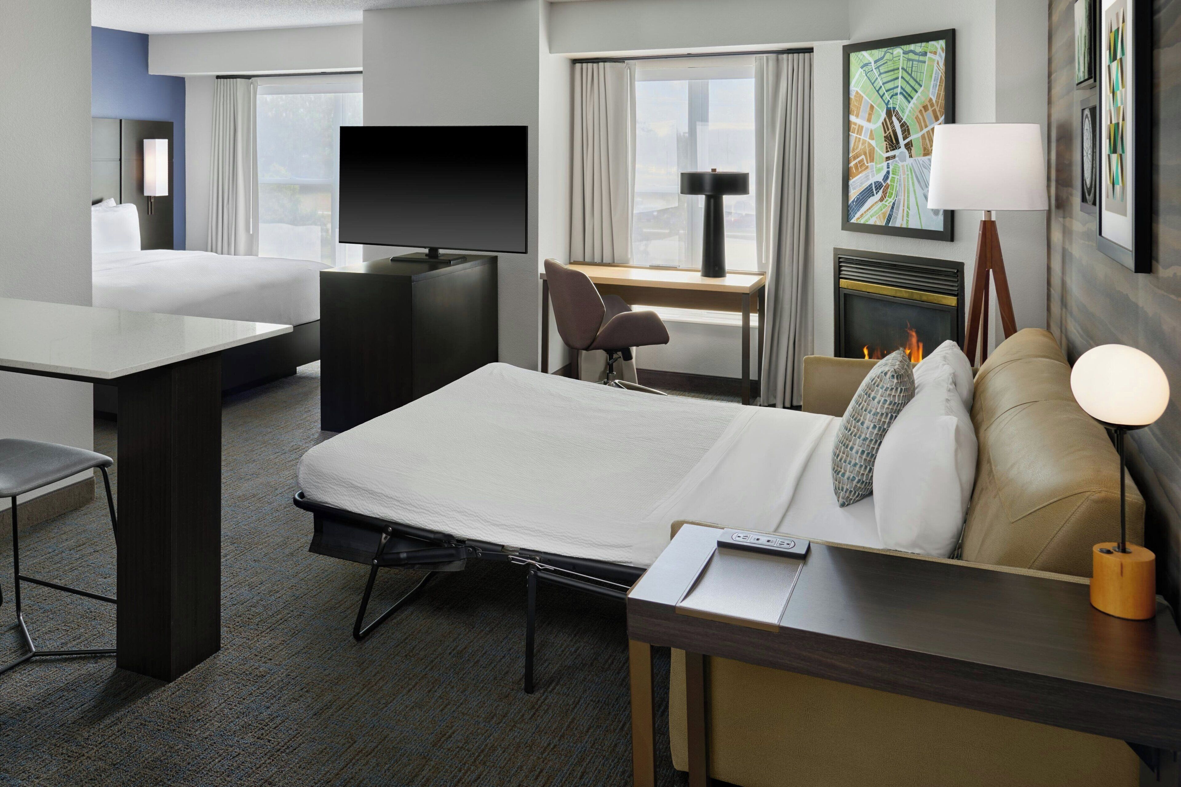 A hotel room with a bed, desk, chair, TV, and a fireplace.