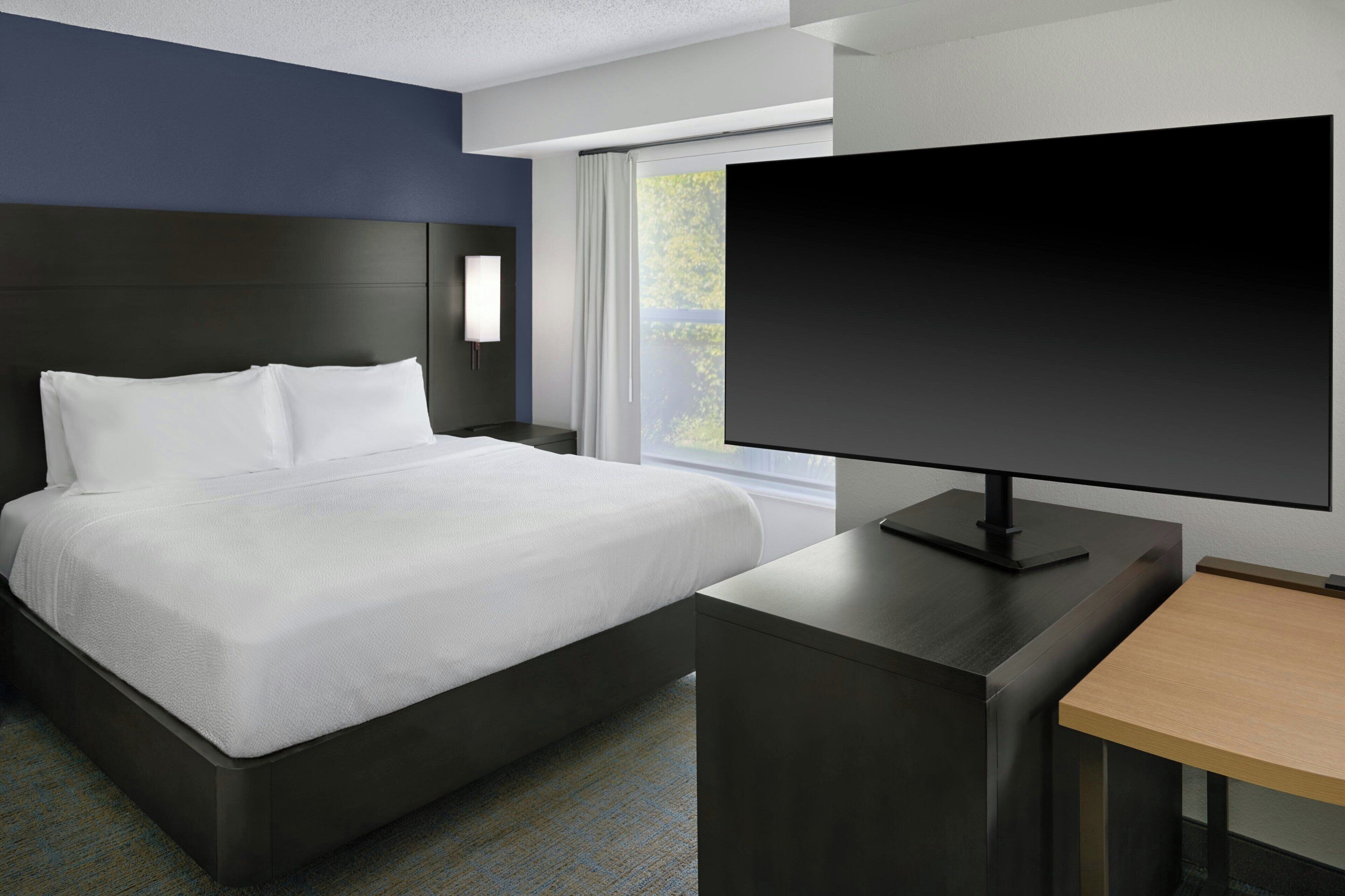 A hotel room with a bed, a desk, a television, and a window with a view of greenery.