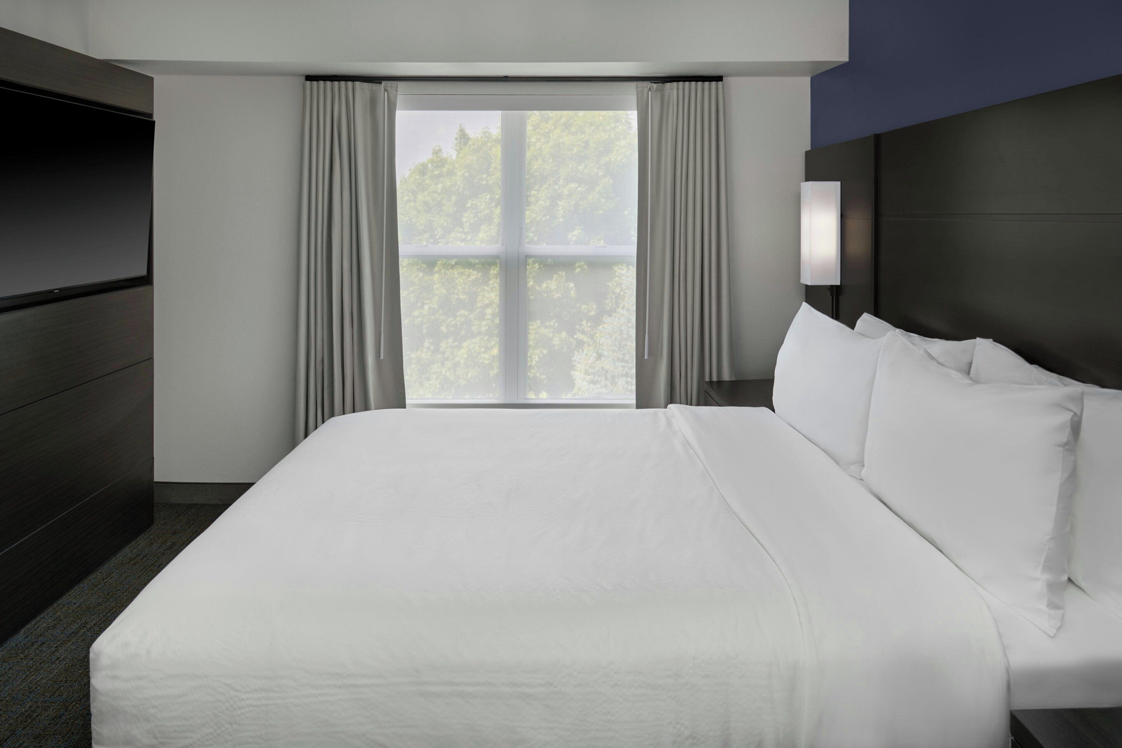 A hotel room with a large bed, a flat-screen TV, a window with curtains, and a nightstand with a lamp.