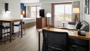 Une chambre d’hôtel comprenant un lit, un bureau avec une chaise, un canapé, une télévision, une lampe et une fenêtre avec des rideaux.