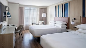 Ein Hotelzimmer mit zwei Betten, einem Schreibtisch, einem Stuhl, einem Fernseher und einem großen Fenster mit Vorhängen.
