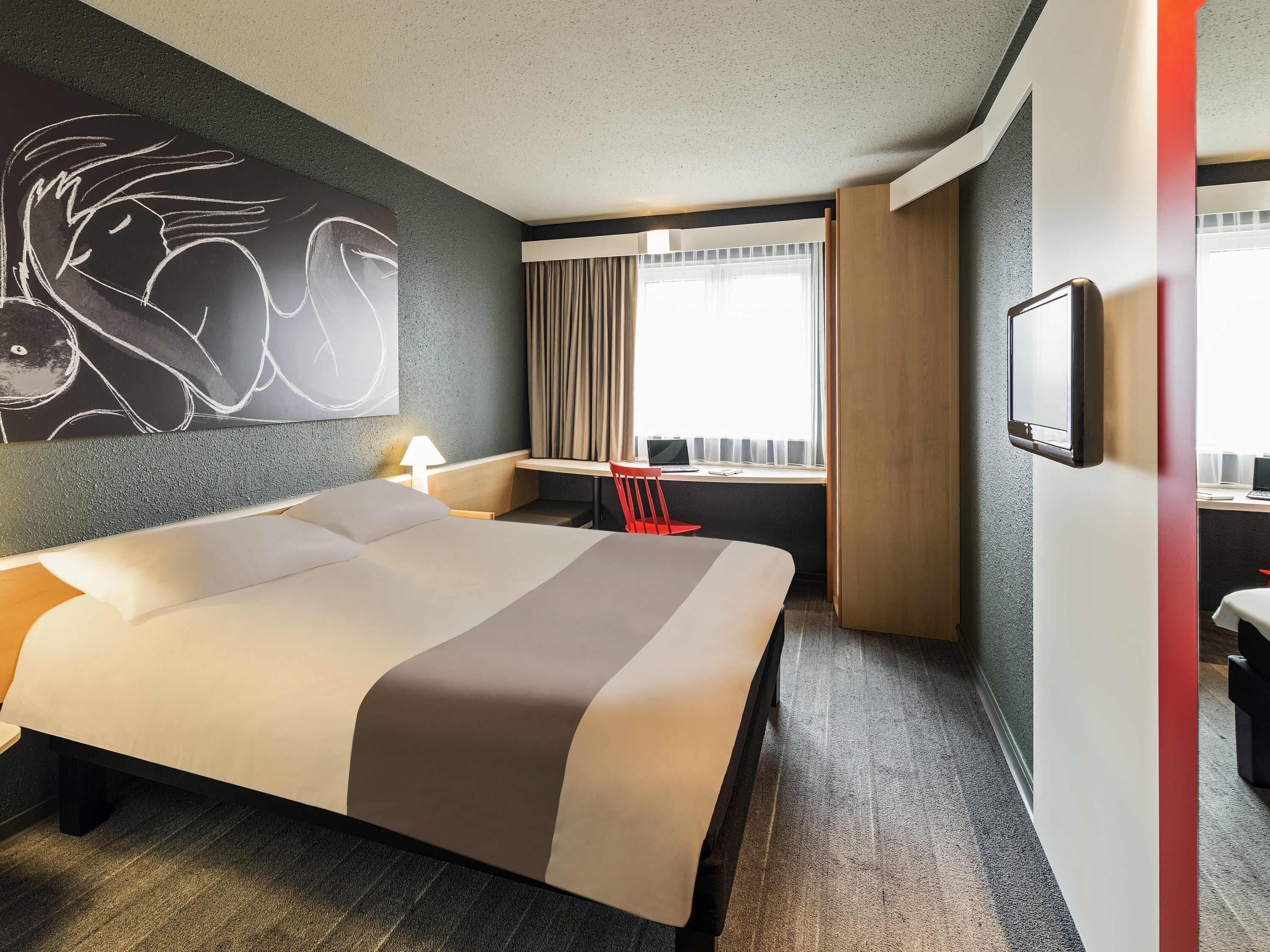A hotel room with a large bed, a desk, a chair, a TV, and a window with curtains.