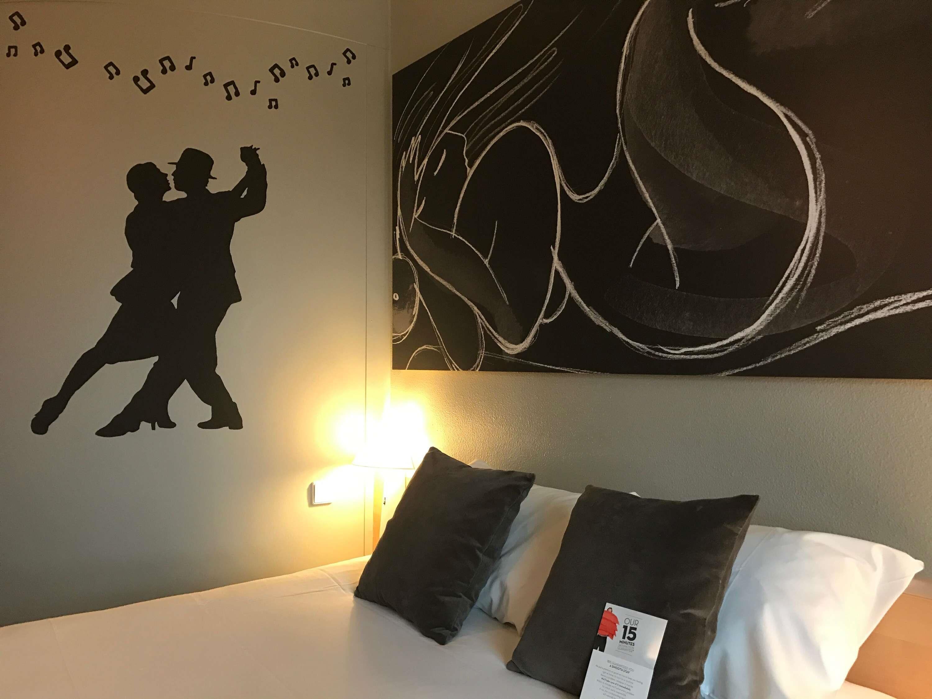 Una habitación de hotel con una cama, almohadas, una obra de arte en la pared de una pareja bailando y un reloj de pared.