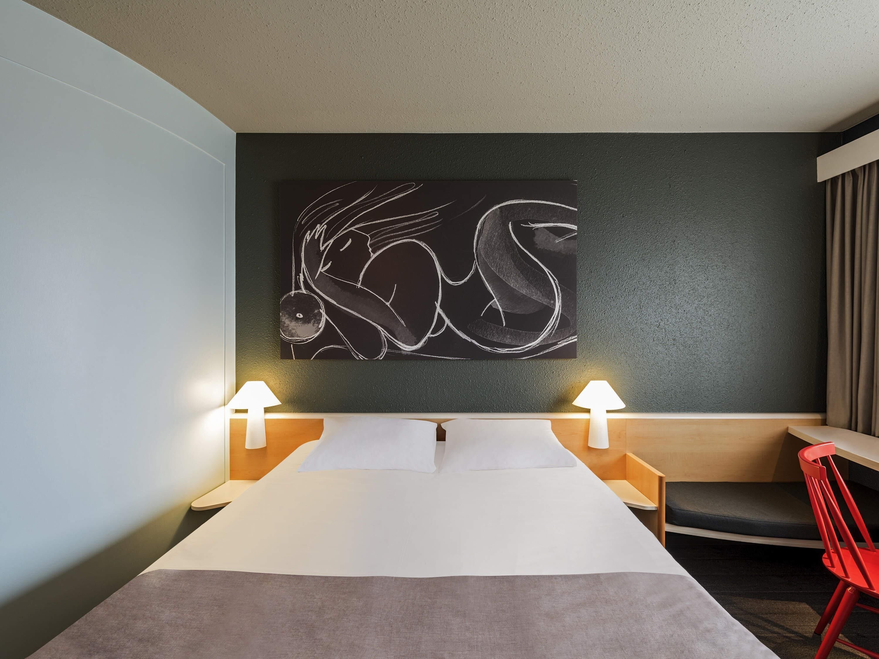Ein Hotelzimmer mit einem Bett, zwei Nachttischlampen, einem roten Sessel, einer dunklen Wand und einem großen abstrakten Kunstwerk.