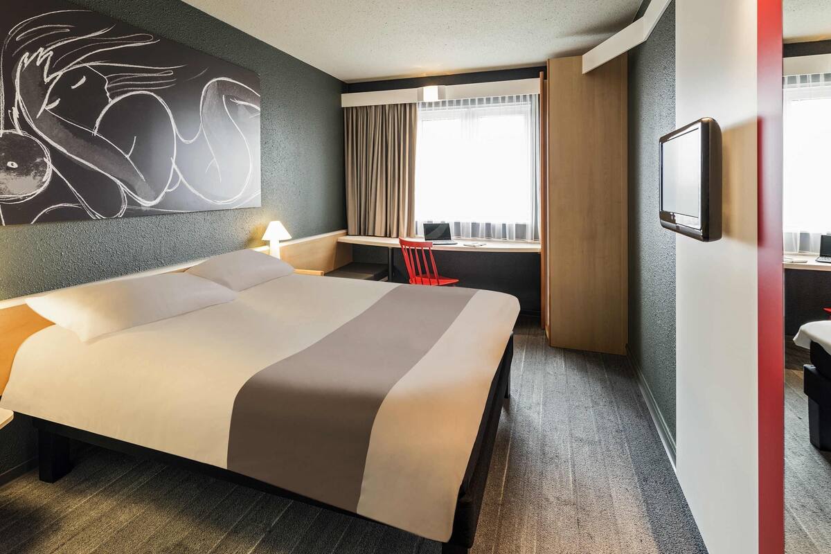 A hotel room with a large bed, a desk with a computer, a chair, a window with curtains, and a wall-mounted TV.