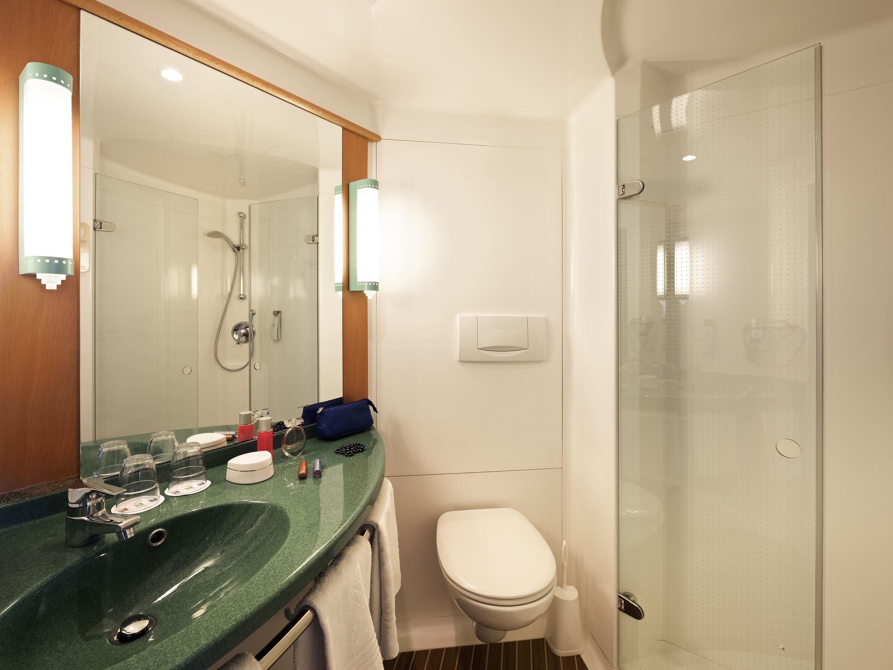 A bathroom with a green sink, a white toilet, a glass shower enclosure, and a mirror with lights.