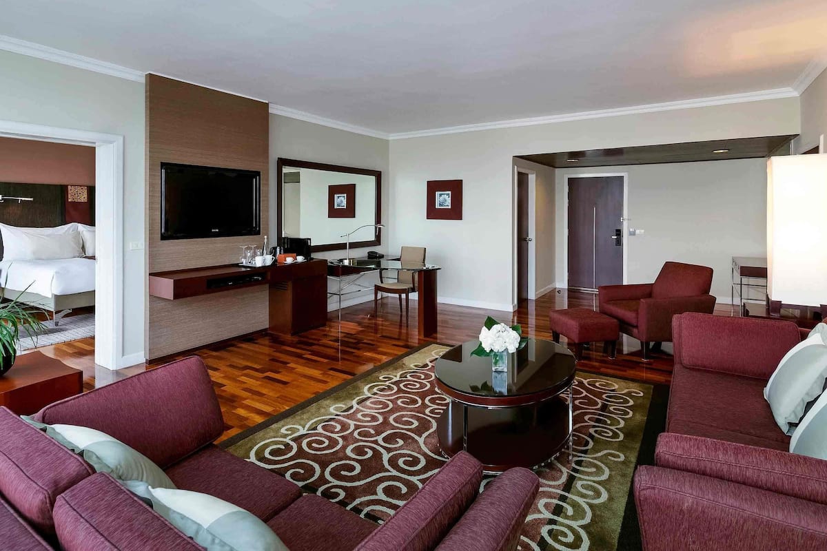 A hotel room with a bed, a seating area with sofas, a coffee table, a TV, a desk with a chair, and a large mirror.