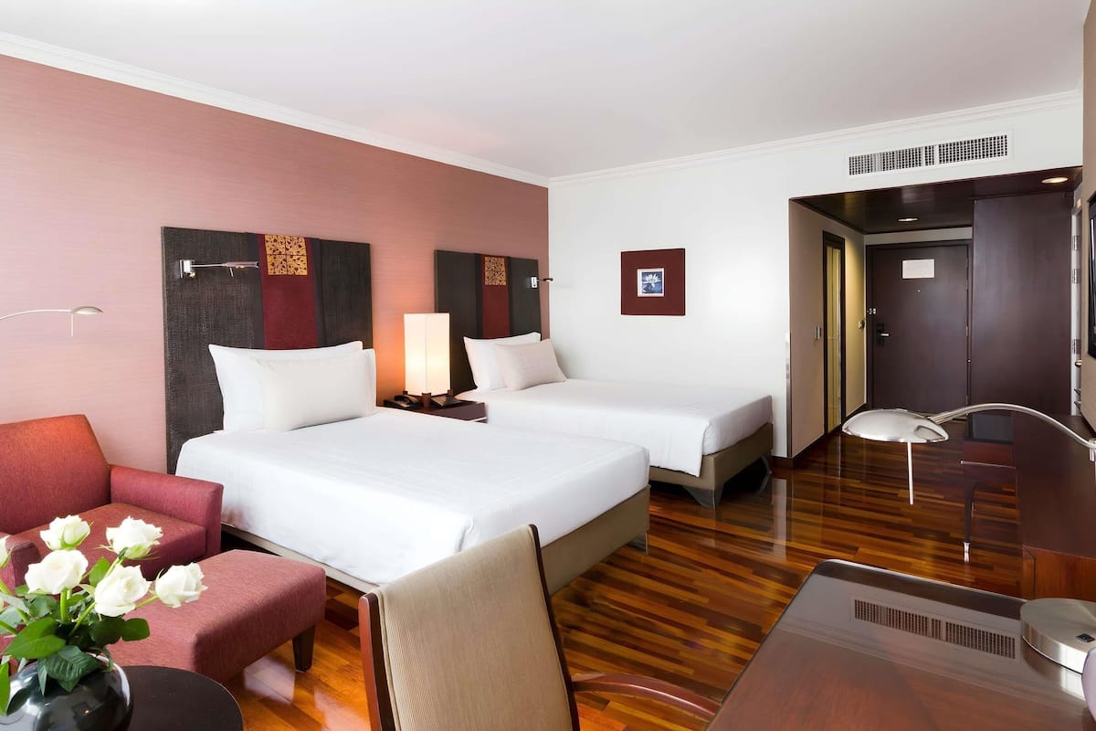 A hotel room with two beds, a desk, a chair, a lamp, and a vase with flowers.