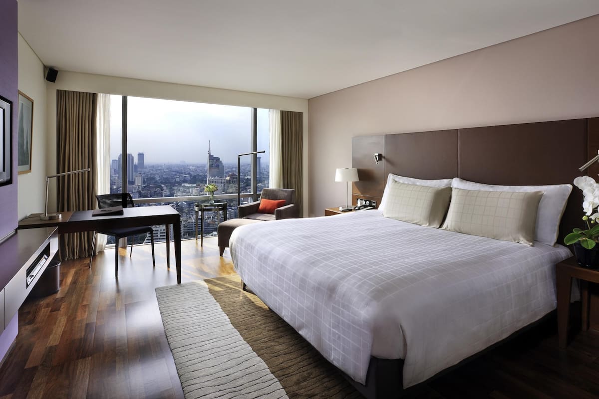 A hotel room with a large bed, a desk, a chair, a nightstand, and a view of a cityscape.