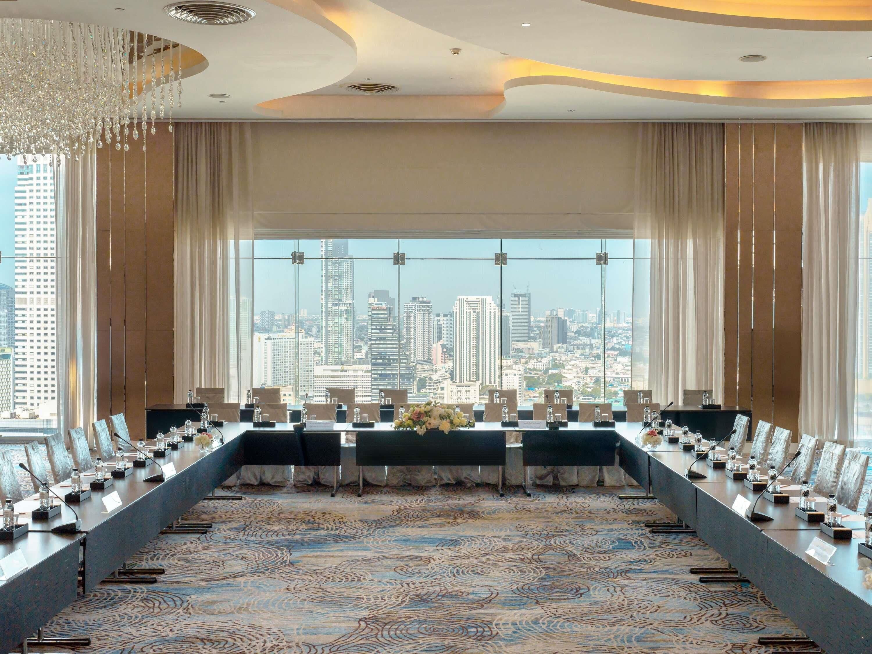 A conference room with a long table, chairs, microphones, and nameplates, with a cityscape view through large windows.