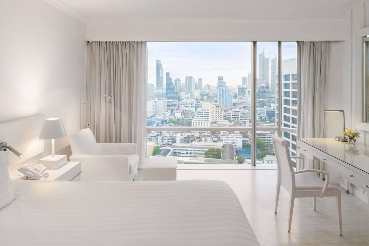 A modern hotel room with a large bed, a desk, a chair, and a view of a cityscape through large windows.