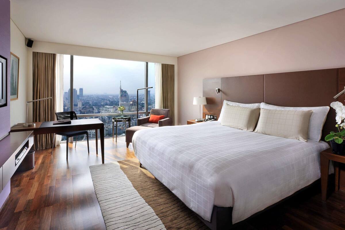 A hotel room with a large bed, a desk, a chair, a nightstand, and a view of a cityscape.