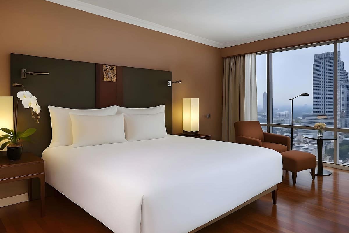A hotel room with a large bed, a chair, a nightstand with a lamp, a potted plant, and a view of a cityscape through large windows.