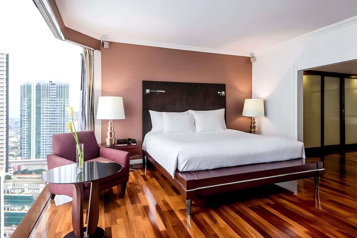 A hotel room with a bed, two bedside tables with lamps, a chair, a small table, and a view of the cityscape.