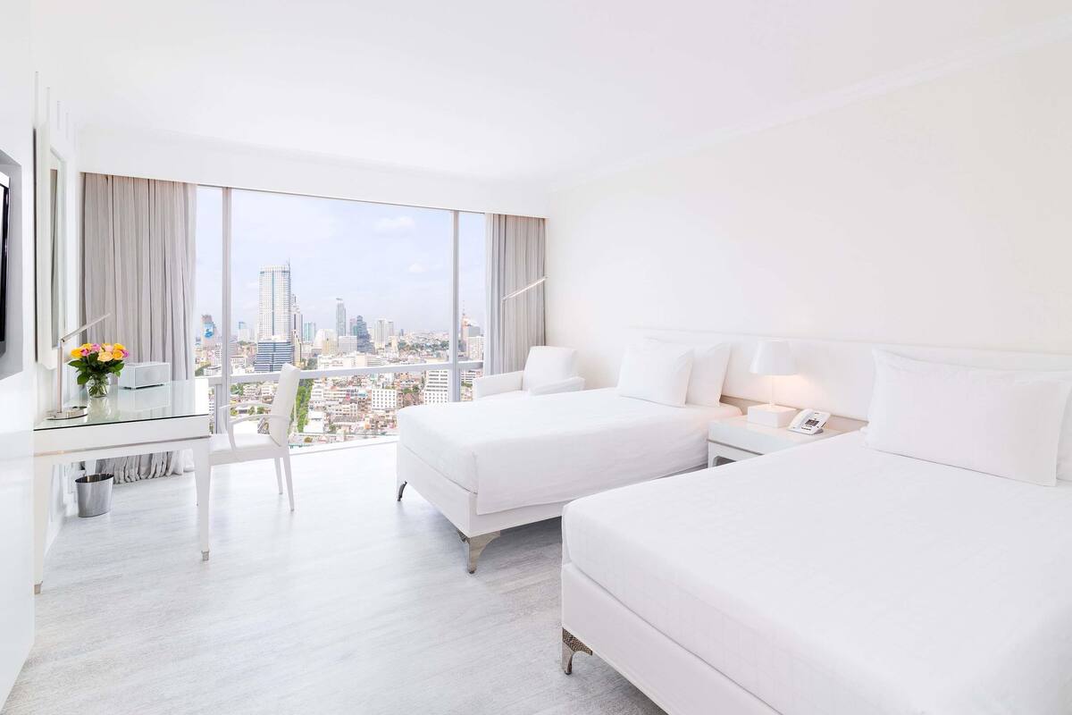 A hotel room with a large window, a bed, a desk with a lamp, and a chair.