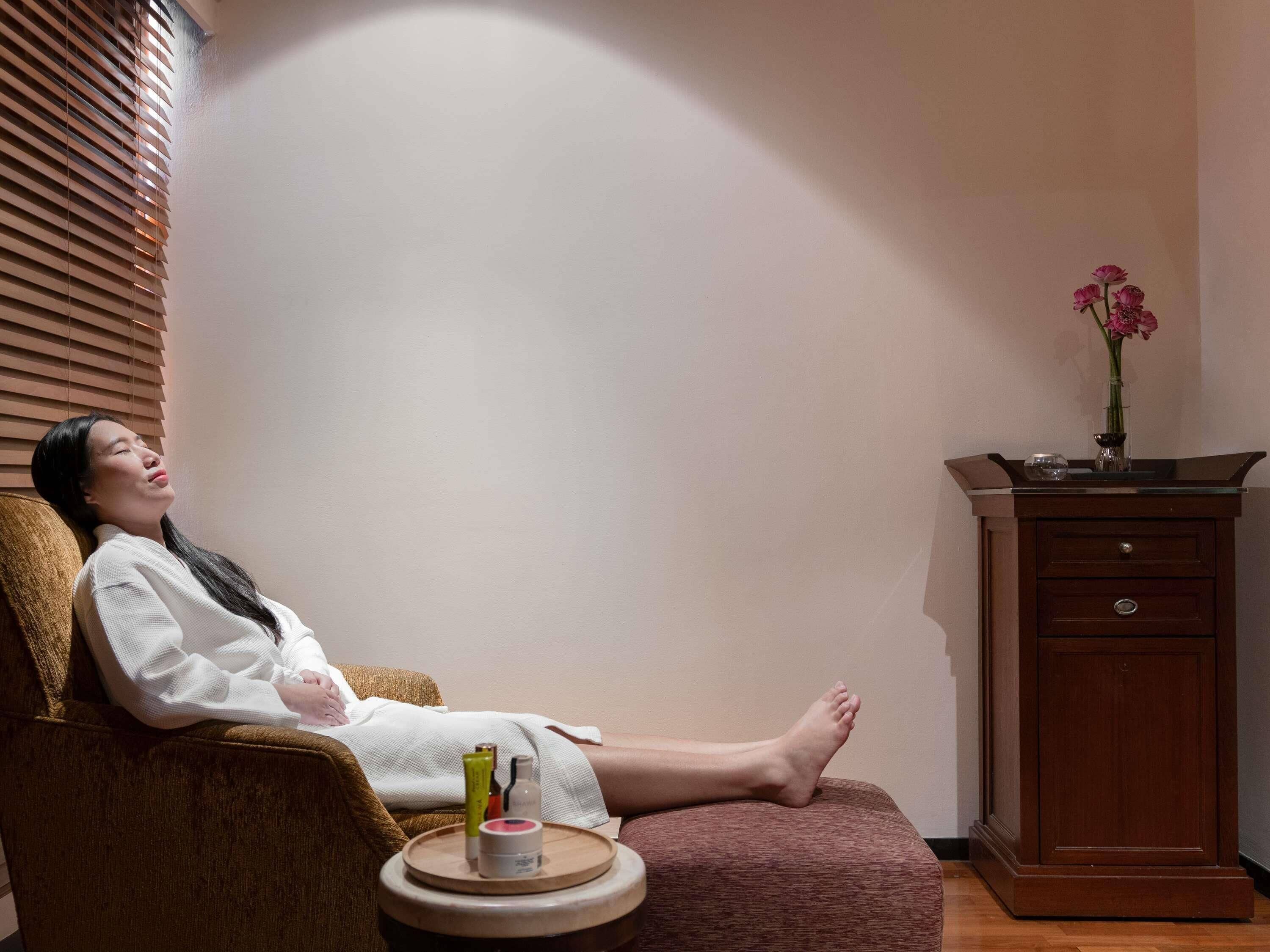 A person relaxing in a chair with a small table beside them containing a bottle, a container, and a cup.