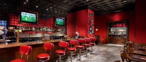 Zona de bar con taburetes rojos, una barra con pantalla de televisión y una pared roja decorada con cuadros.
