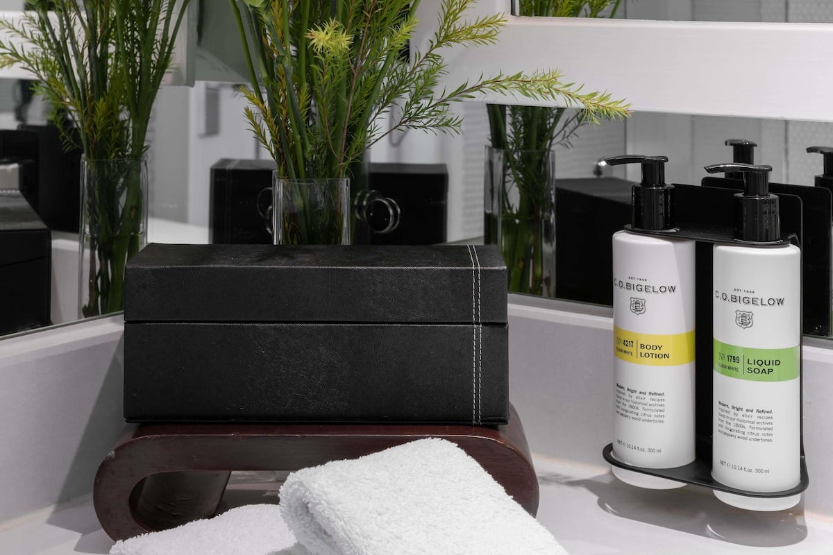 A bathroom with a black toiletry bag, two bottles of C.O. Bigelow body lotion and liquid soap, a white towel, and a vase with green plants.
