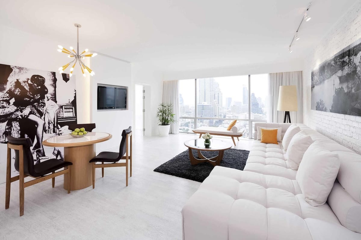 A modern living room with a white sofa, a dining table with chairs, a TV mounted on the wall, and a large window with a city view.