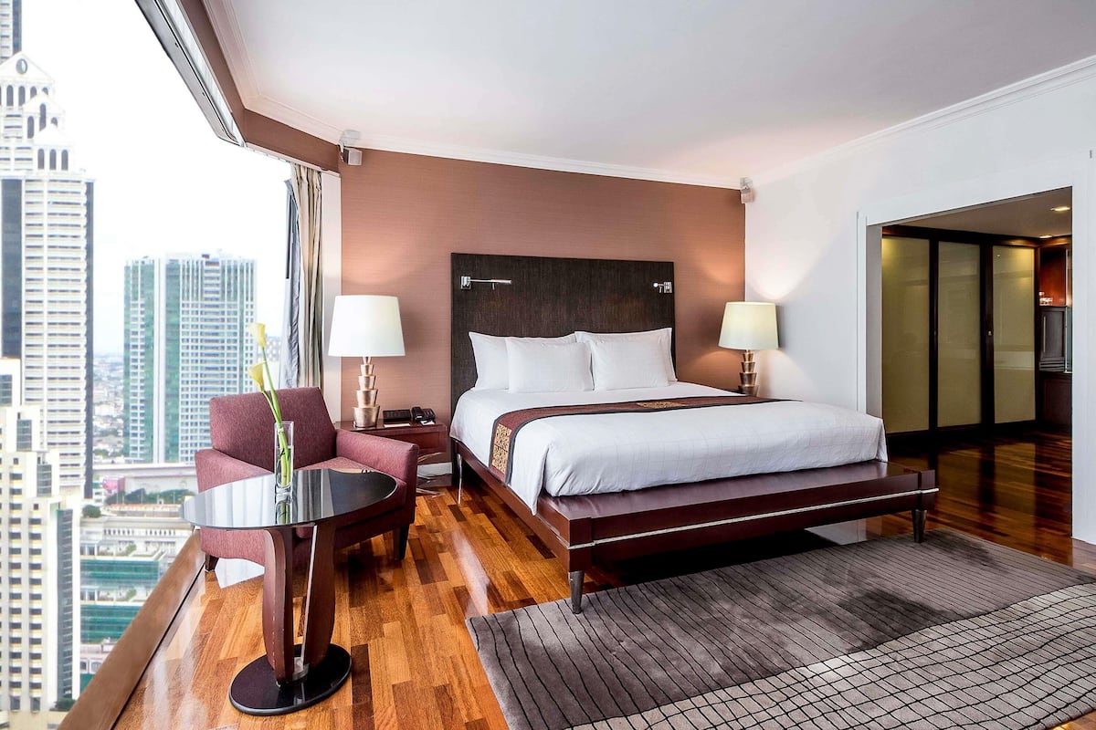 A hotel room with a bed, bedside tables, a chair, a small table, and a view of a cityscape.