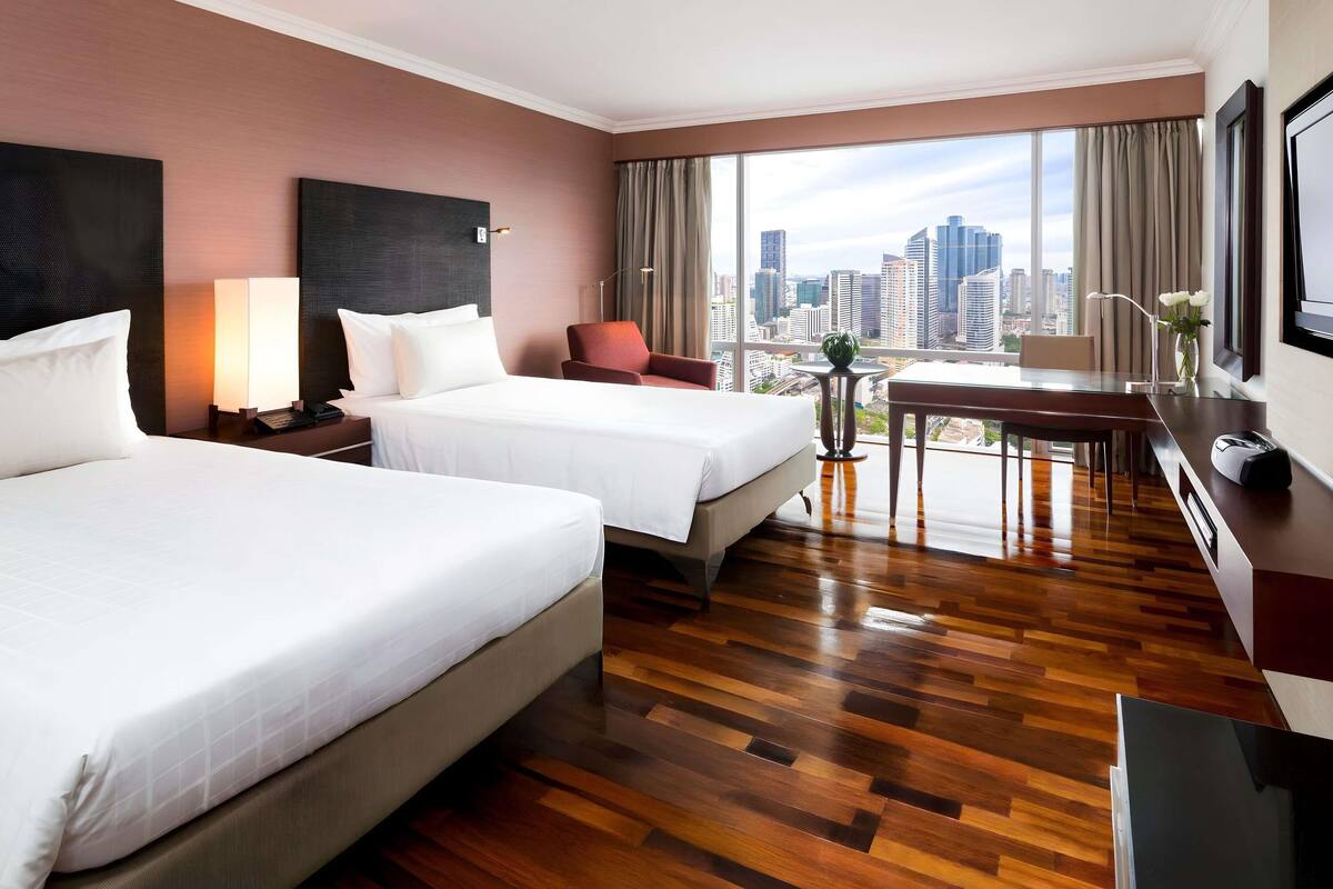 A hotel room with two beds, a desk, a chair, a lamp, and a view of the cityscape.