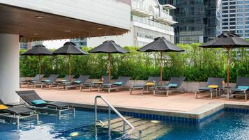 Zona junto a la piscina con sillones de descanso y sombrillas, rodeada de vegetación y edificios modernos.