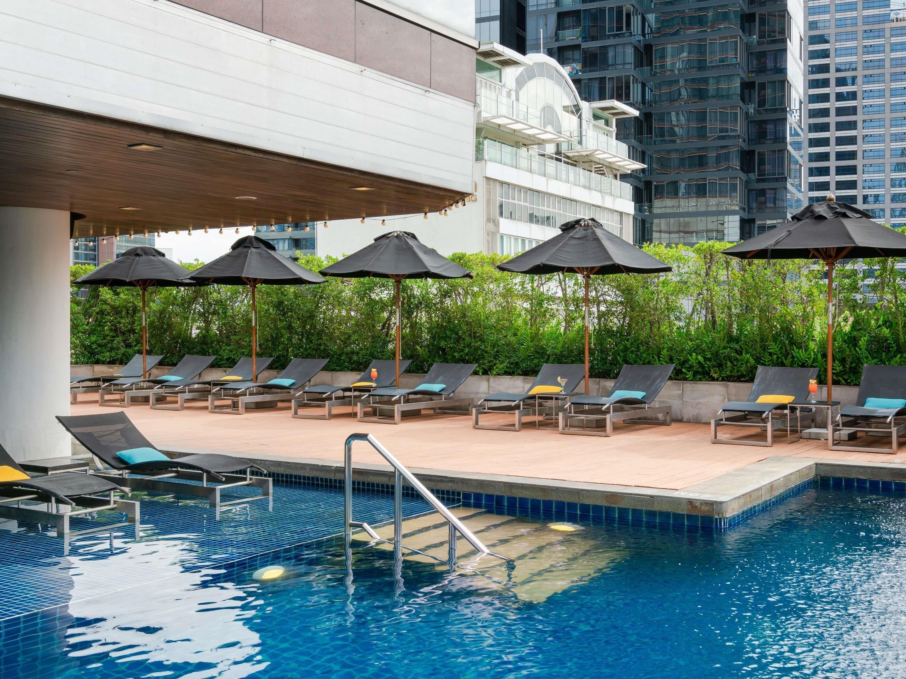 A rooftop pool area with lounge chairs and umbrellas, surrounded by greenery and modern buildings.