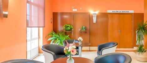 A modern waiting area with orange walls, a round table with chairs, and a sign reading "SALÓN LA ENCINA".