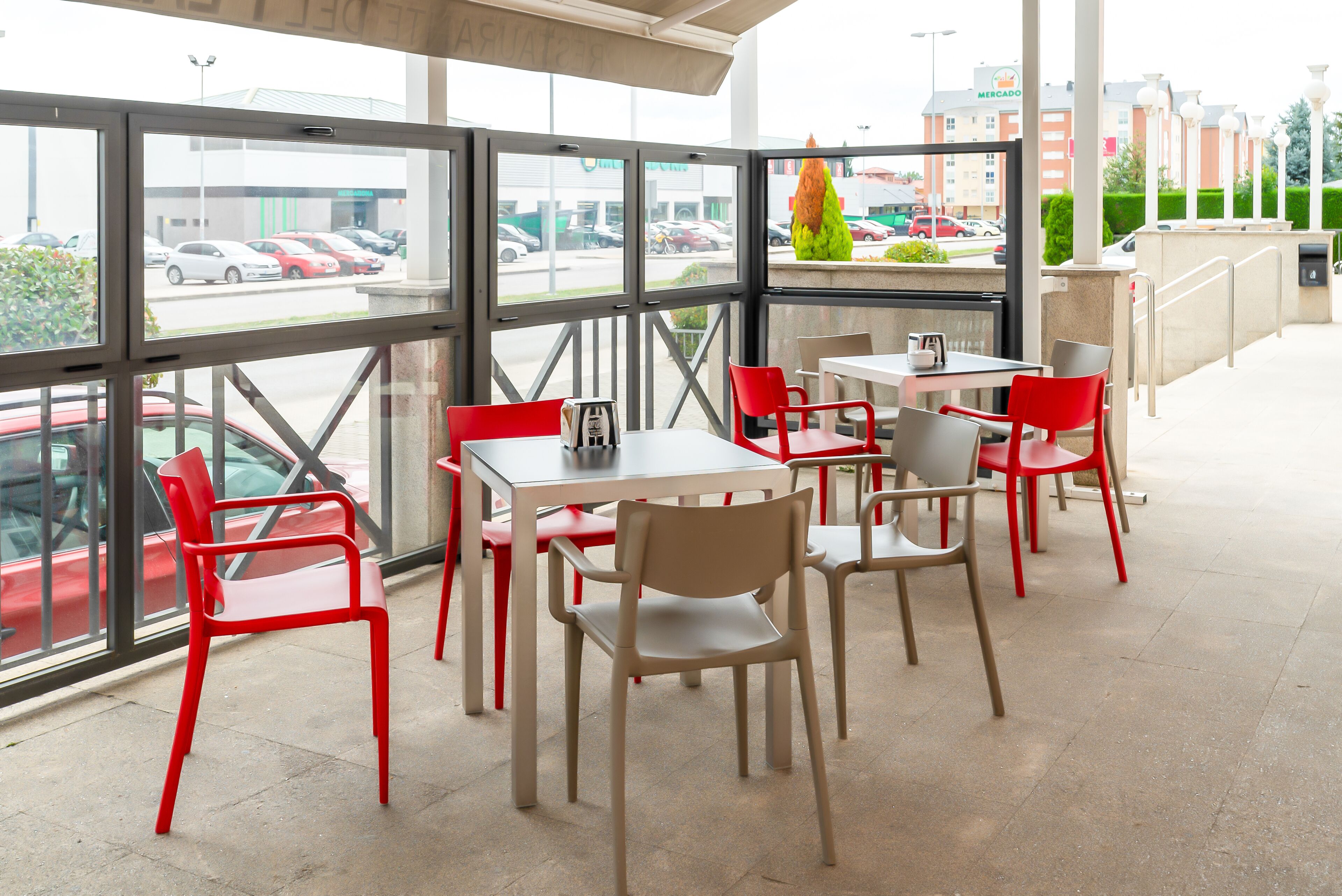 Un restaurante con amplios ventanales, sillas rojas y beige, y vistas a coches aparcados y edificios exteriores.