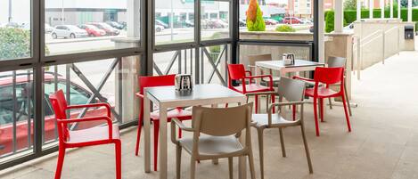Un restaurante con amplios ventanales, sillas rojas y beige, y vistas a coches aparcados y edificios exteriores.