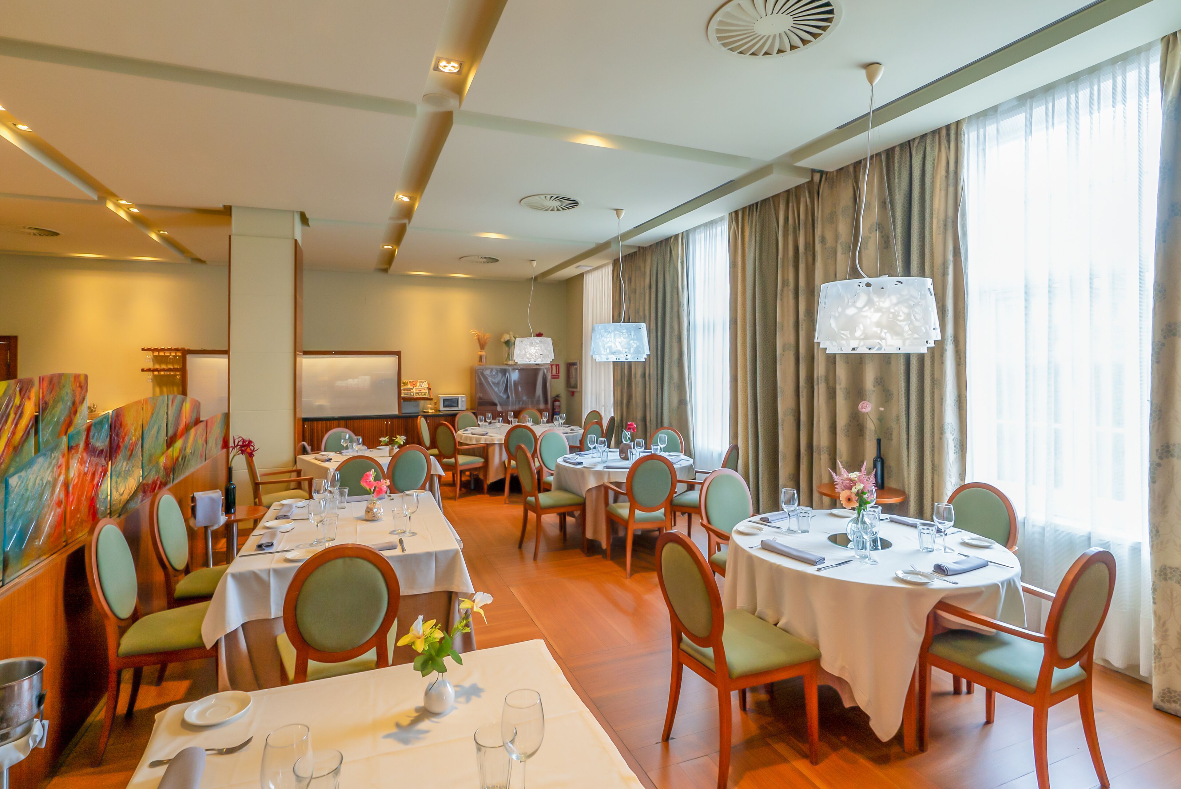 Un comedor de hotel con varias mesas redondas, sillas tapizadas de verde y manteles blancos.