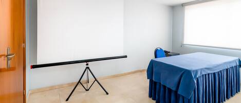 Un salón de conferencias con una pantalla de proyección, una mesa con mantel azul y un ventanal.