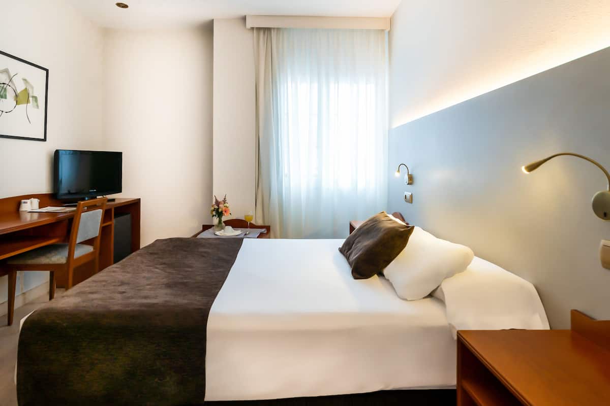Habitación de hotel con cama, escritorio con computadora, silla, televisor y ventana con cortinas.