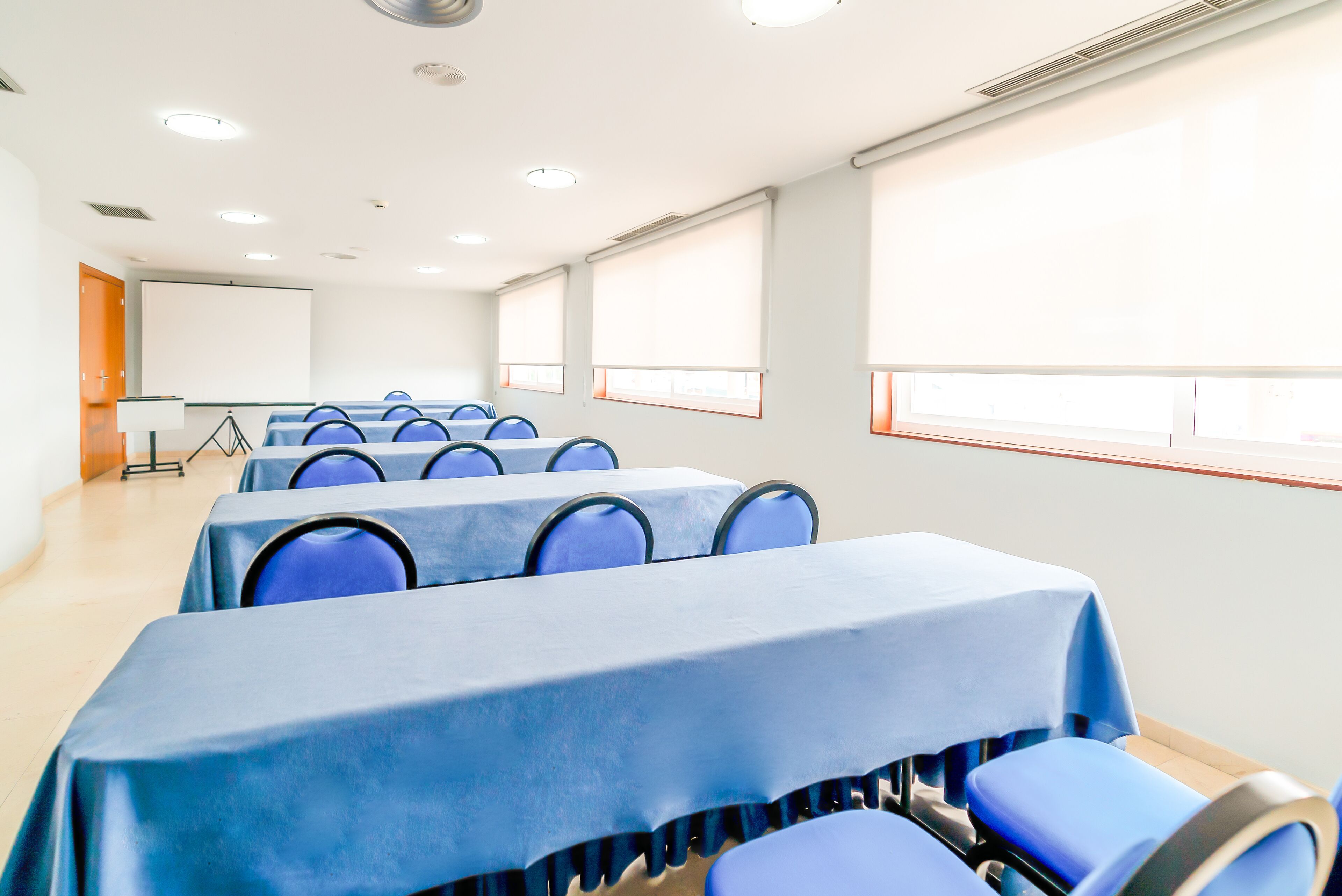 Une salle de conférence avec des rangées de tables et de chaises, un écran de projection et de grandes fenêtres.