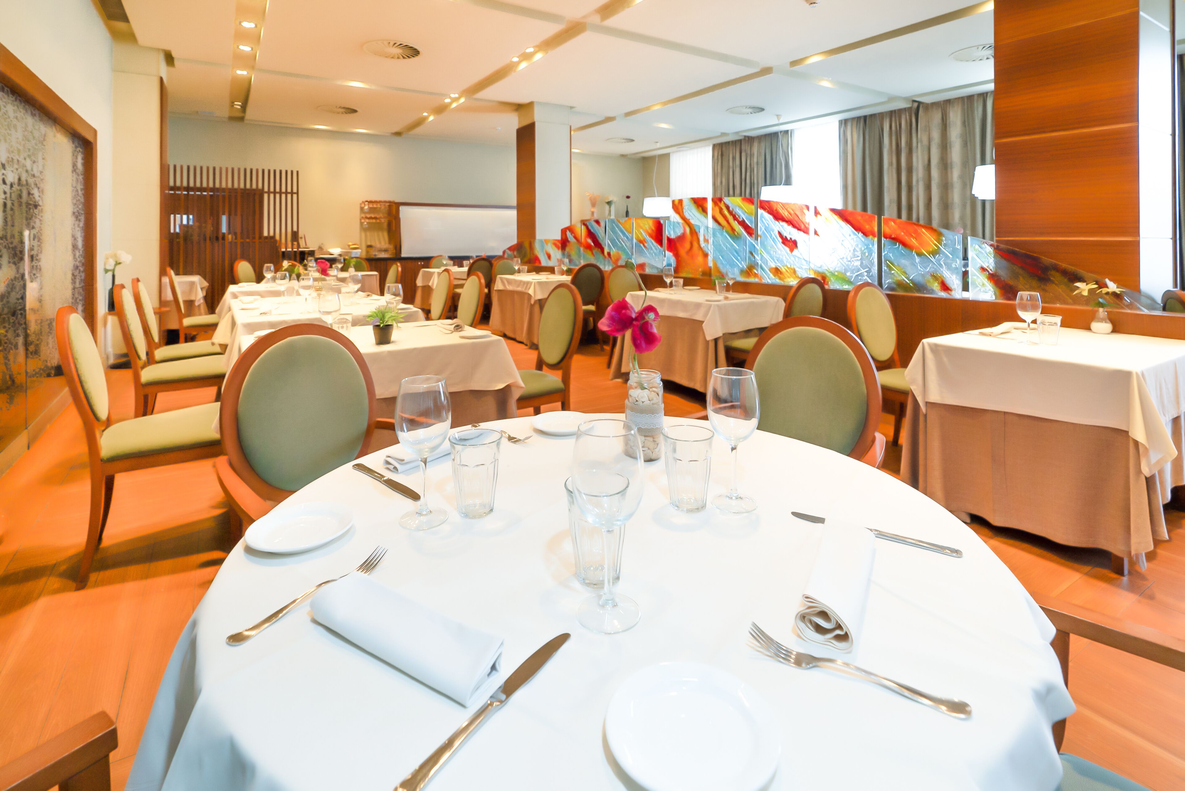 Un comedor de hotel con mesas redondas dispuestas para comer, sillas y un gran cuadro abstracto en la pared.