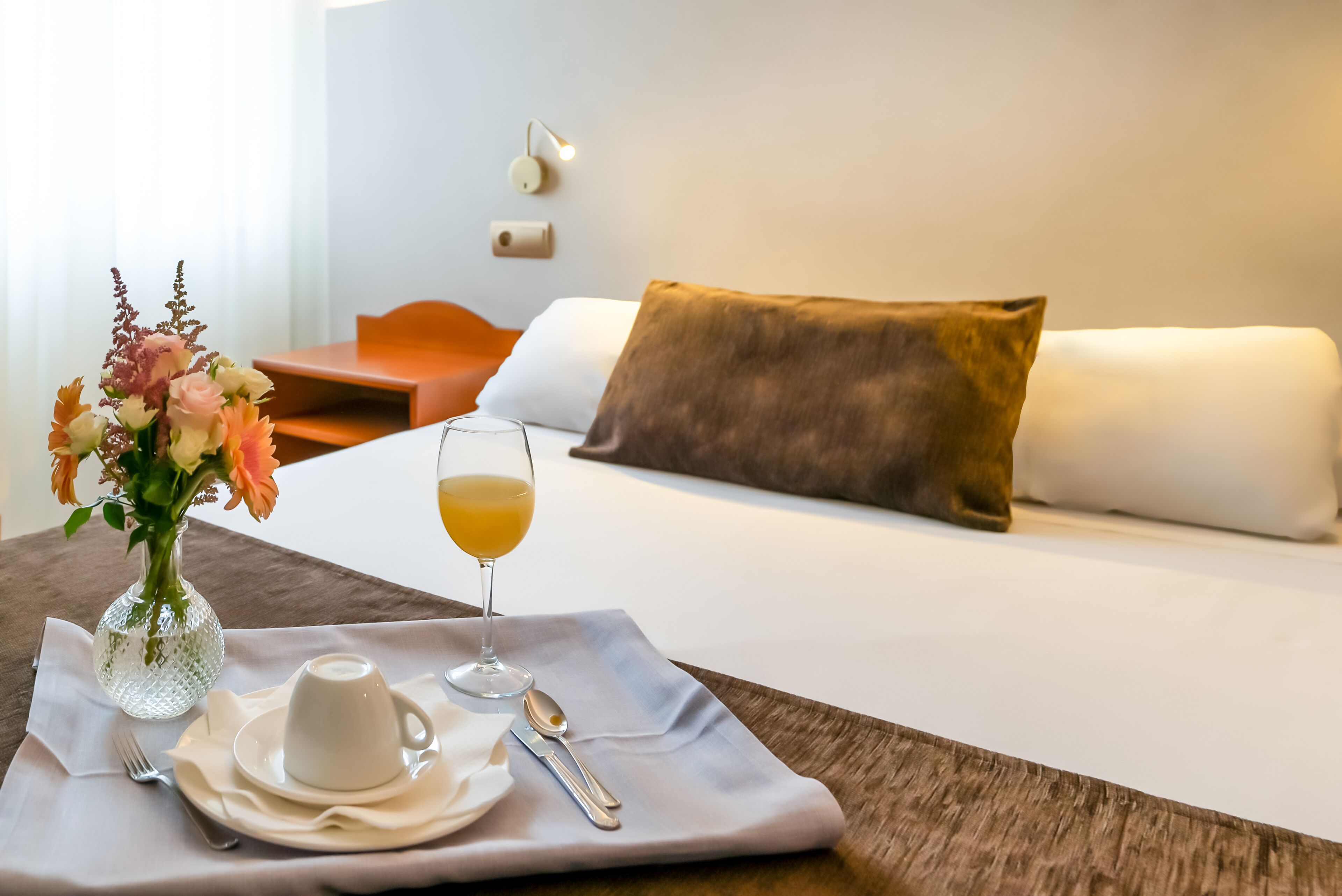 A neatly arranged breakfast table with a vase of flowers, a glass of orange juice, a white cup, and a spoon on a white napkin.