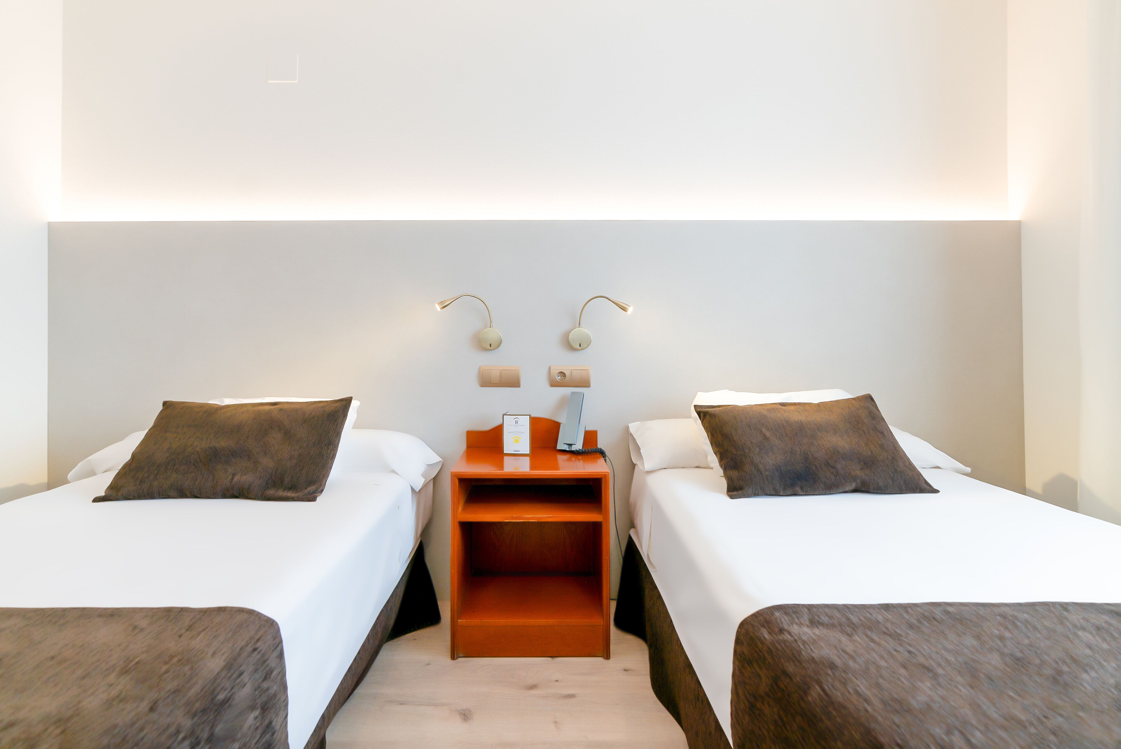 Habitación de hotel con dos camas individuales, una mesita de noche con teléfono y lámpara, y una lámpara de pared.