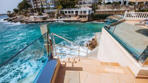 Un hotel frente al mar con una piscina infinita de fondo de cristal, una escalera que lleva a la piscina y vistas al océano y edificios cercanos.