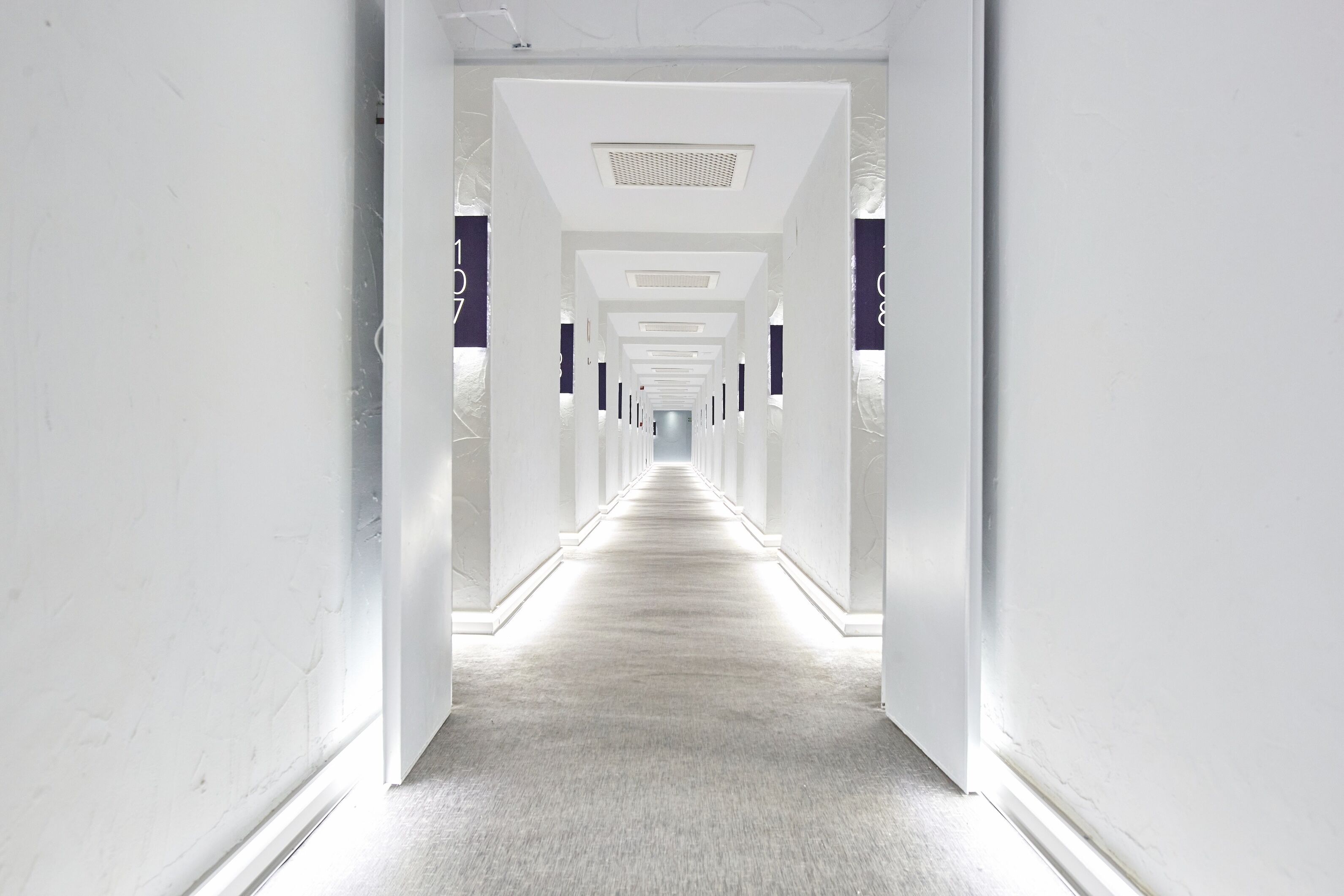 Un pasillo largo y estrecho con paredes blancas y suelo alfombrado, que conduce a una habitación luminosa al final.