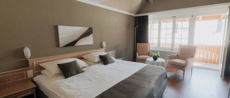 A hotel room with a large bed, a small round table, a chair, and a balcony with a view.
